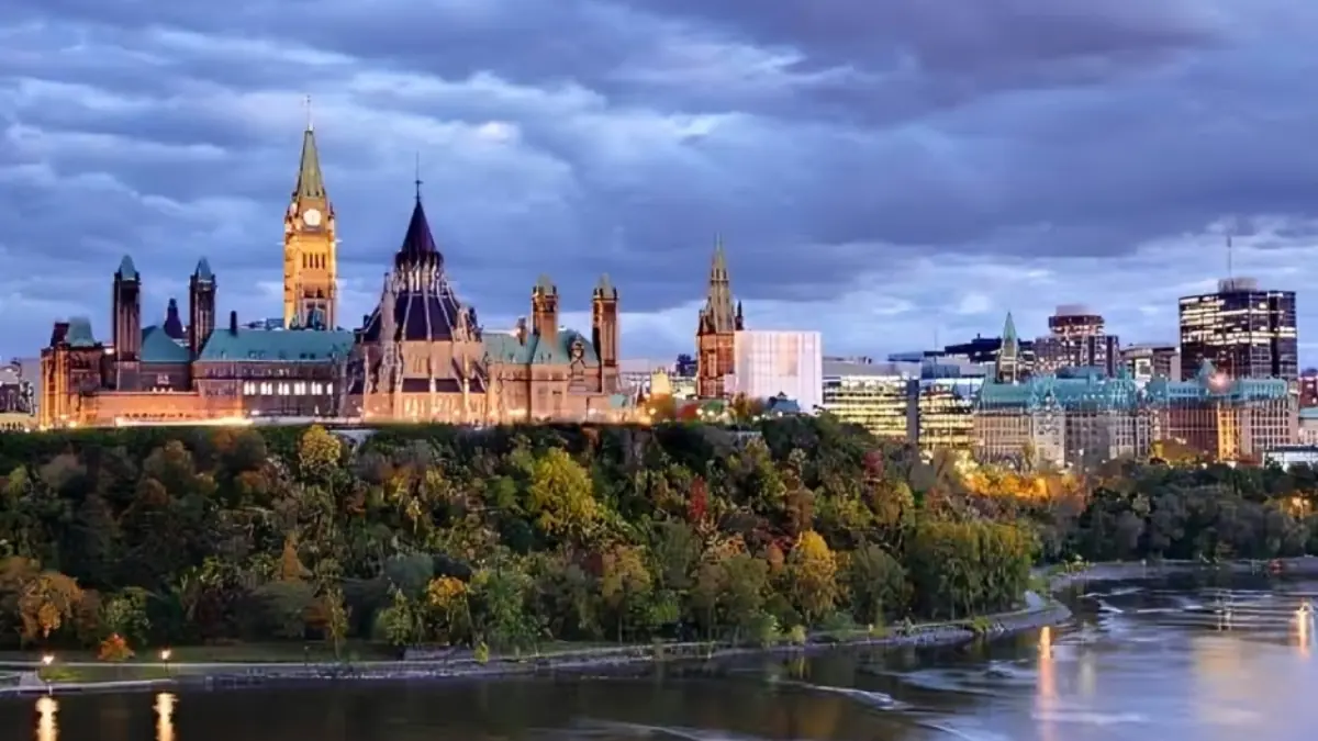 Parliament Hill Ottawa - THEWILL NEWS MEDIA Ottawa Parliament Hill at dusk, with lit buildings, trees in foreground and the river in front.