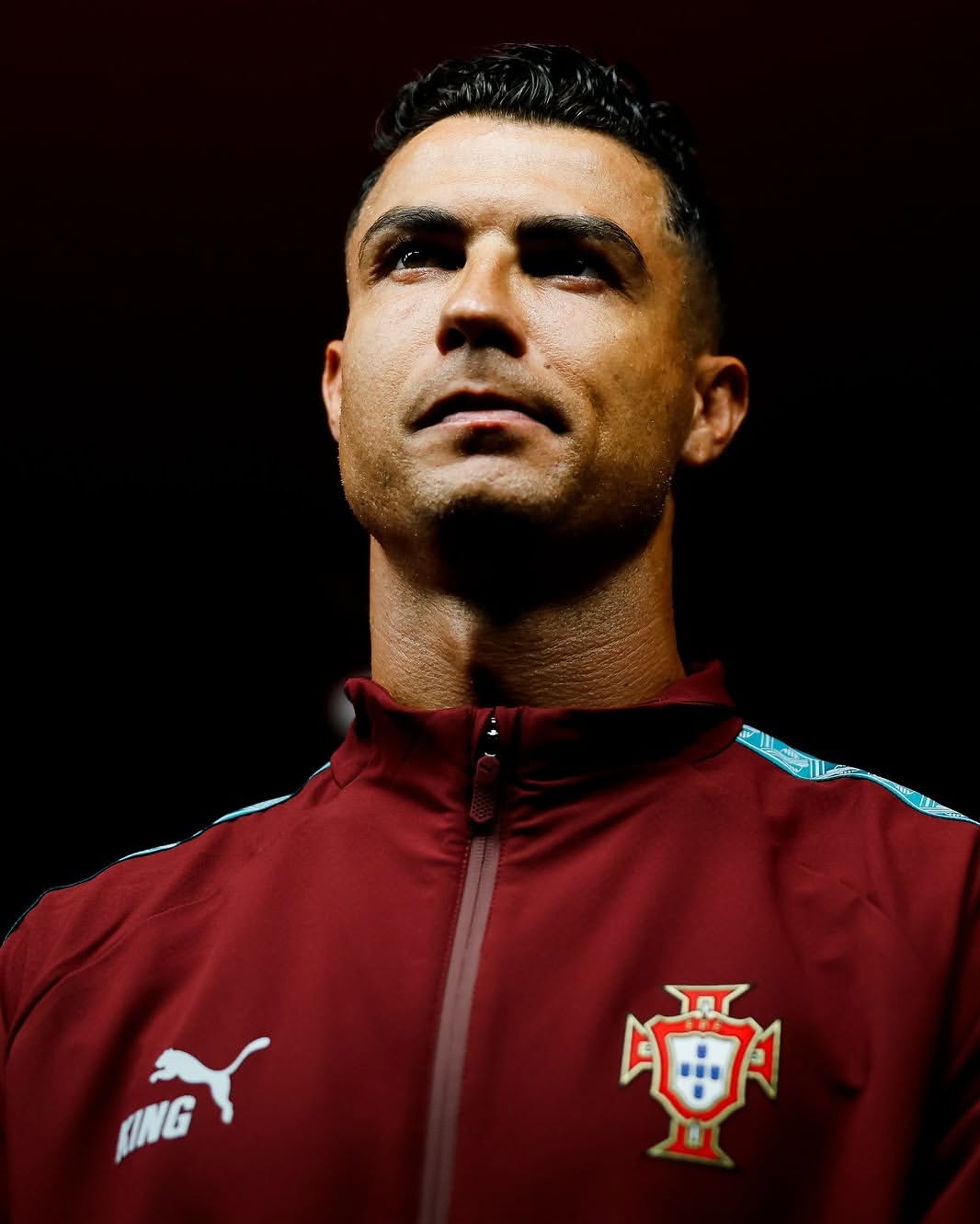 Portugal Sempre - THEWILL NEWS MEDIA Close-up portrait of a man in a maroon jacket with the Portugal crest, looking upward against a dark background.