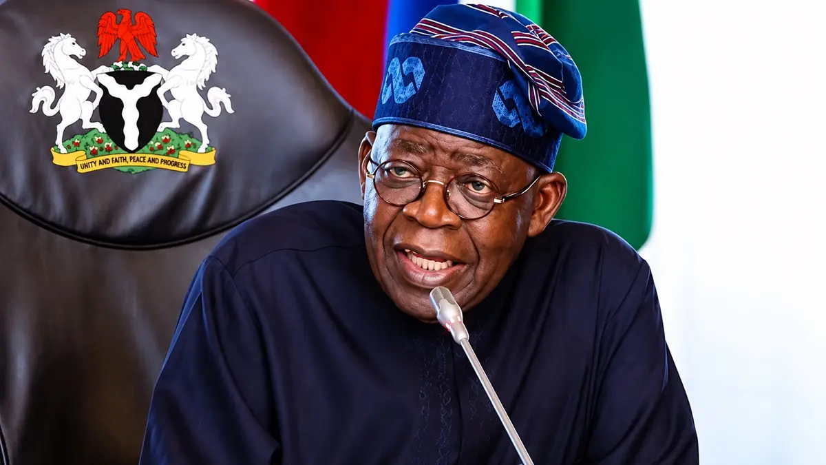 President Bola Tinubu - THEWILL NEWS MEDIA Smiling man in navy traditional attire and cap speaks into a desk microphone at a formal meeting, with a government emblem on the chair behind him.
