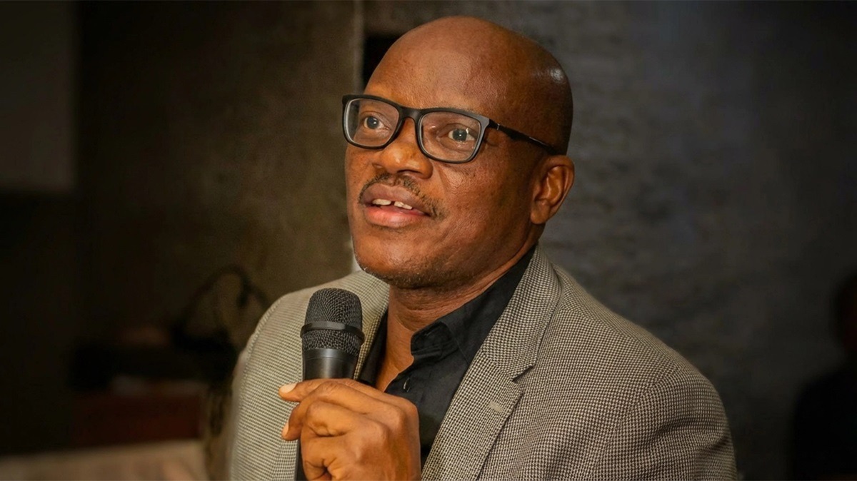Prof Christopher Piwuna - THEWILL NEWS MEDIA Man wearing glasses and a gray blazer speaks into a handheld microphone.