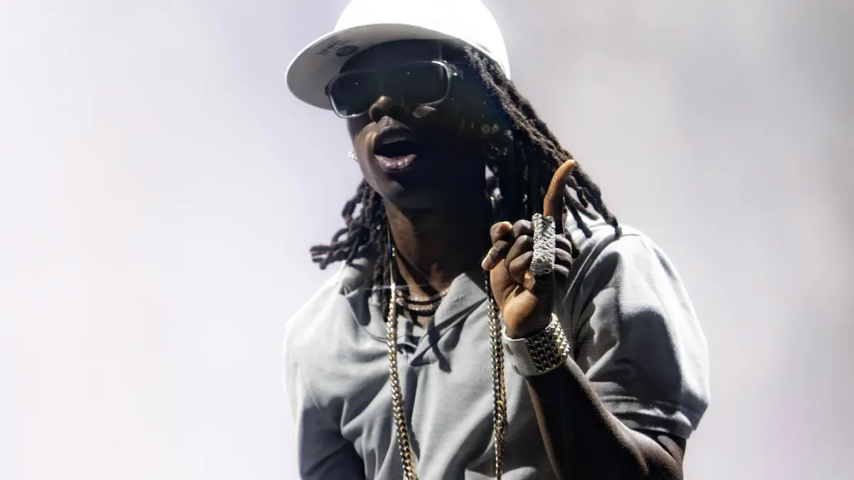 Rema - THEWILL NEWS MEDIA Male hip-hop artist performing on stage in a white cap and sunglasses, gesturing with a raised finger and chains visible.