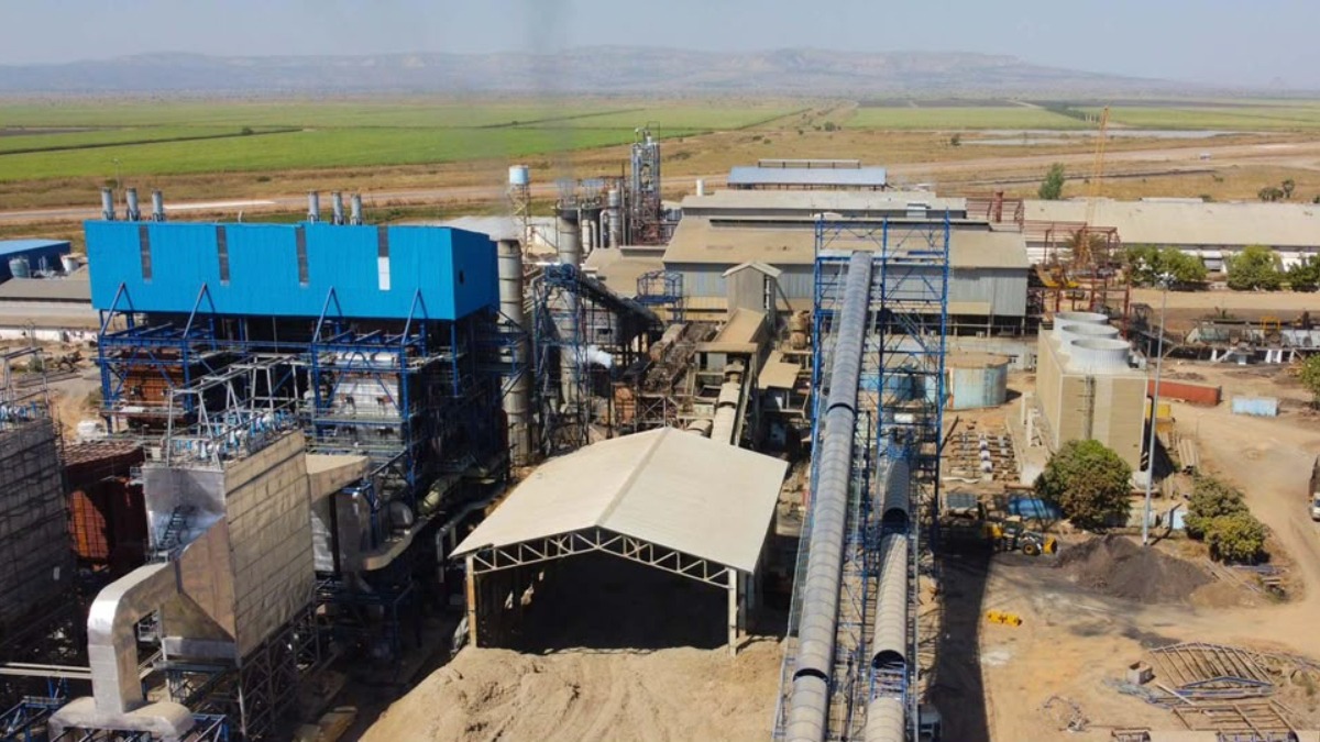Representation of expansion - THEWILL NEWS MEDIA Industrial processing plant with blue buildings, long conveyor belts, and storage tanks in a dry, rural area