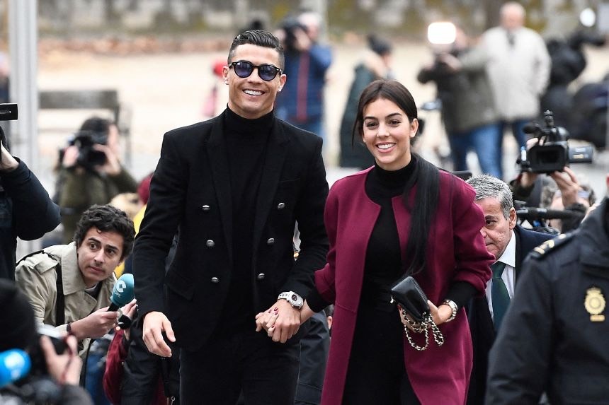 Ronaldo and Georgina - THEWILL NEWS MEDIA Man in a black double-breasted suit and sunglasses walks hand in hand with a woman in a burgundy coat as photographers crowd around them.