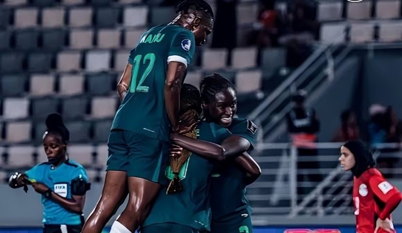 Super Falcons - THEWILL NEWS MEDIA Female soccer players in teal uniforms celebrate a goal, multiple players group-hugging on the field as a teammate jumps on top in a stadium.