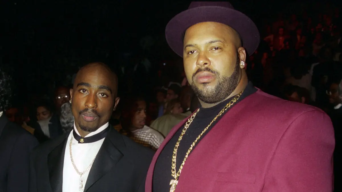 Tupac Shakur and Marion Suge - THEWILL NEWS MEDIA Two men posing at a nighttime event; one in a black suit with white shirt and gold chain, the other in a burgundy blazer and hat with a gold chain.