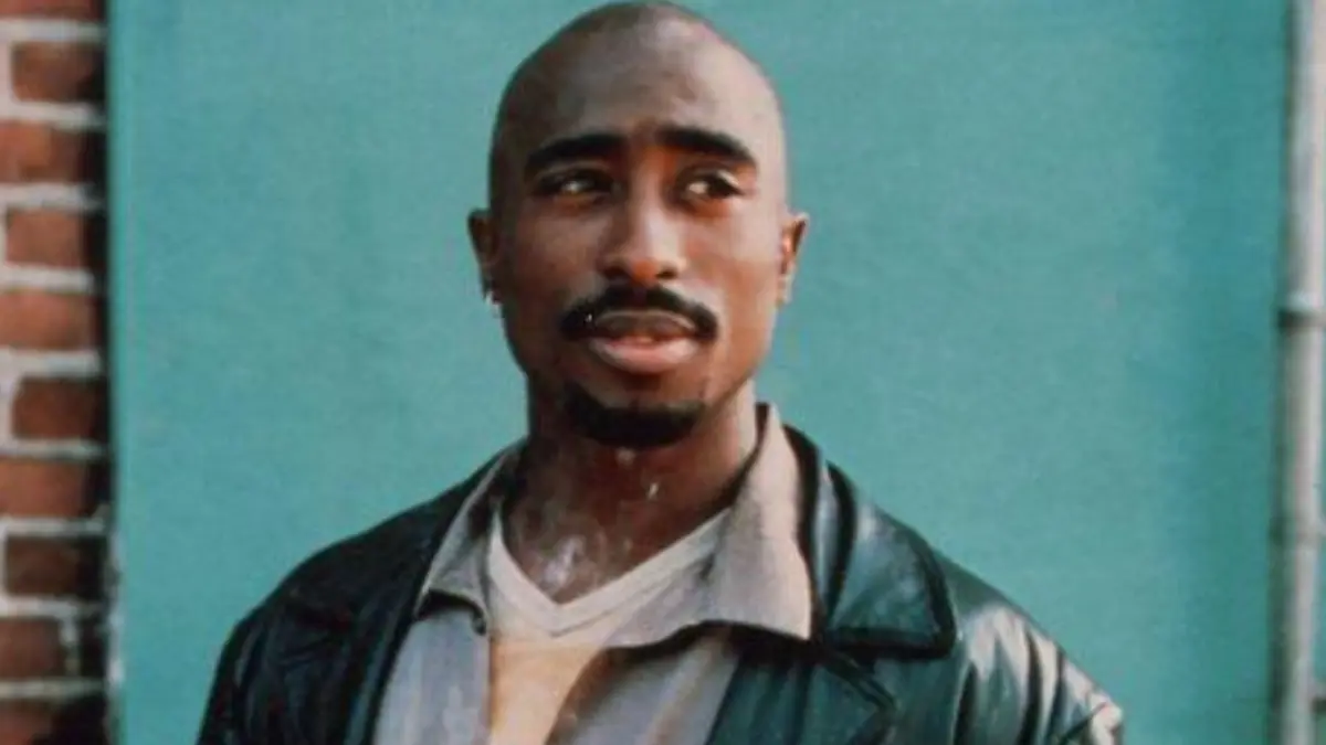 Tupac - THEWILL NEWS MEDIA A man with a shaved head and mustache wearing a dark leather jacket, standing against a teal wall with brick edge on the left.