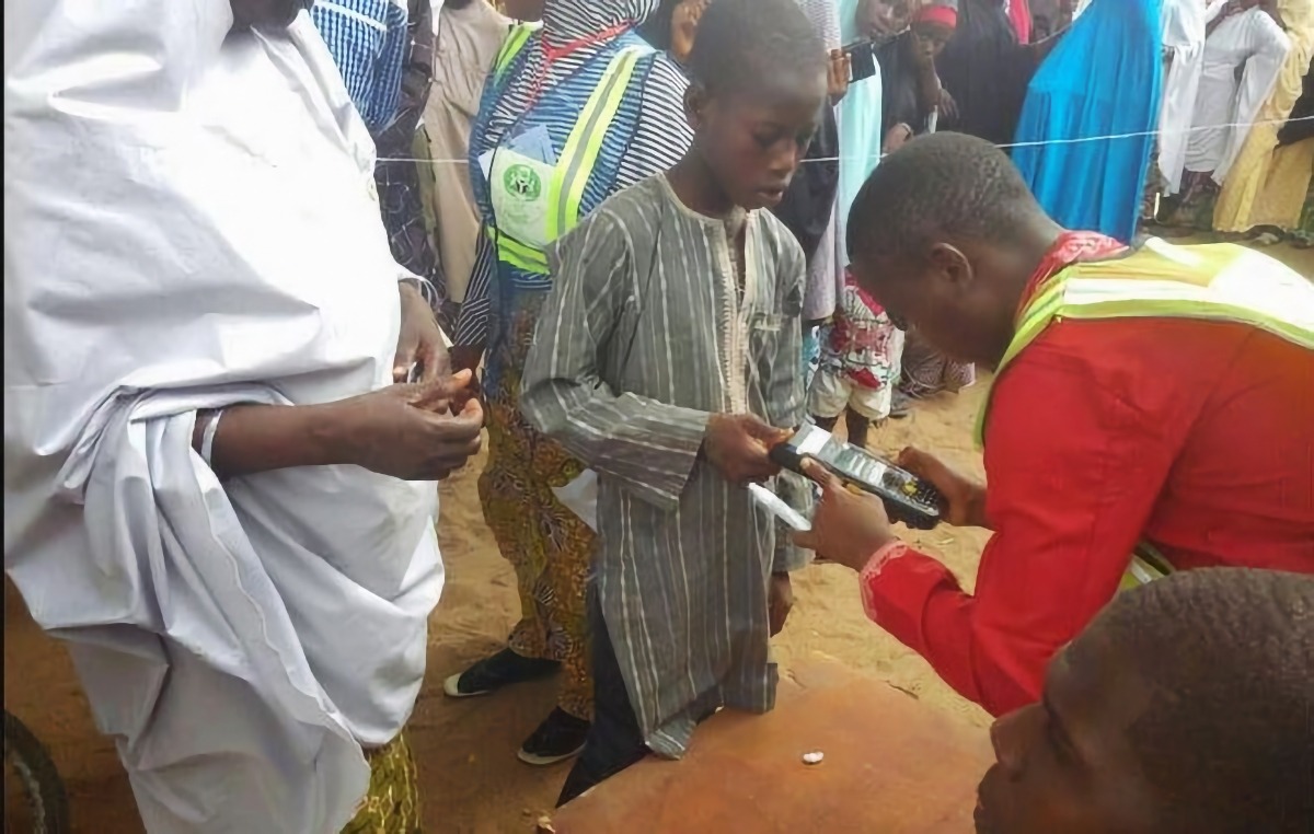 Underage-voting-1 - THEWILL NEWS MEDIA Aid worker in a red uniform with a reflective vest hands a device to a young boy in a striped tunic in a crowded outdoor setting.
