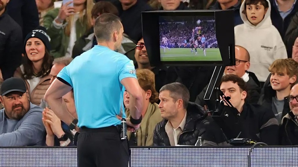 VAR - THEWILL NEWS MEDIA Referee in a blue shirt facing a packed stadium, crowd watching as a monitor shows a football match.