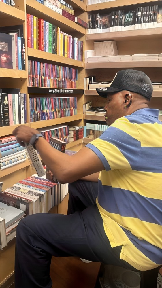 Vice President Kashim Shettima - THEWILL NEWS MEDIA Man wearing a blue and yellow striped polo sits on a stool, browsing books in a bookstore.