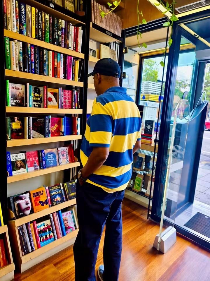 Vice President Kashim Shettima 2 - THEWILL NEWS MEDIA Man in a yellow and blue striped polo browses books on a wooden shelf inside a bookstore.