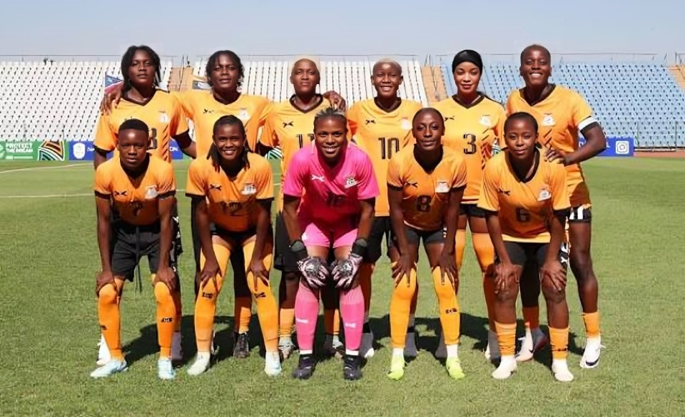 Zambia team Copper Queens - THEWILL NEWS MEDIA Women’s soccer team posing for a group photo on the field, goalkeeper in pink center, all in orange uniforms and numbered jerseys.