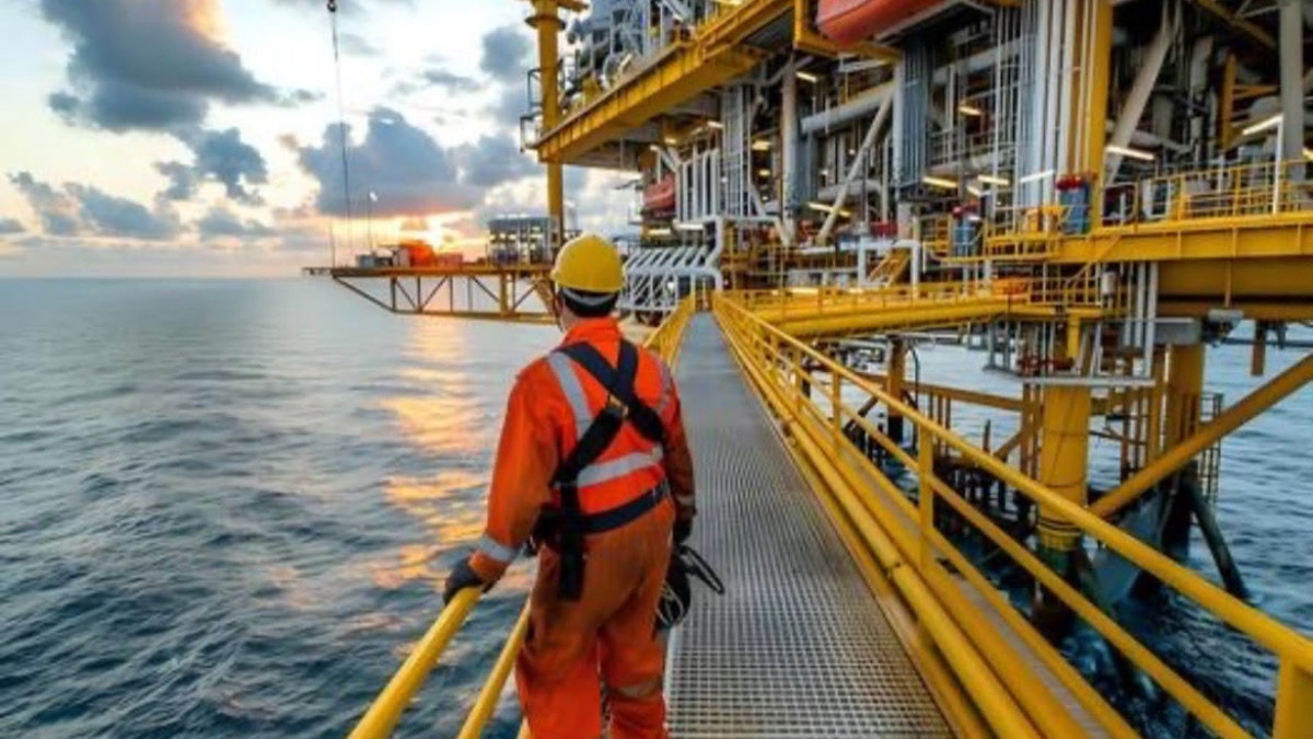 offshore engineer - THEWILL NEWS MEDIA Worker in orange coveralls and yellow hard hat walks the metal catwalk of an offshore oil platform at sunset.