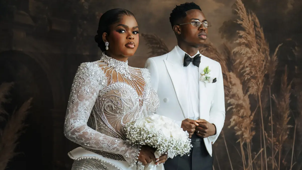 peller jarvis white wedding - THEWILL NEWS MEDIA Bride in an embellished sheer lace gown holds a white bouquet, standing beside a groom in a white tuxedo with a black bow tie.
