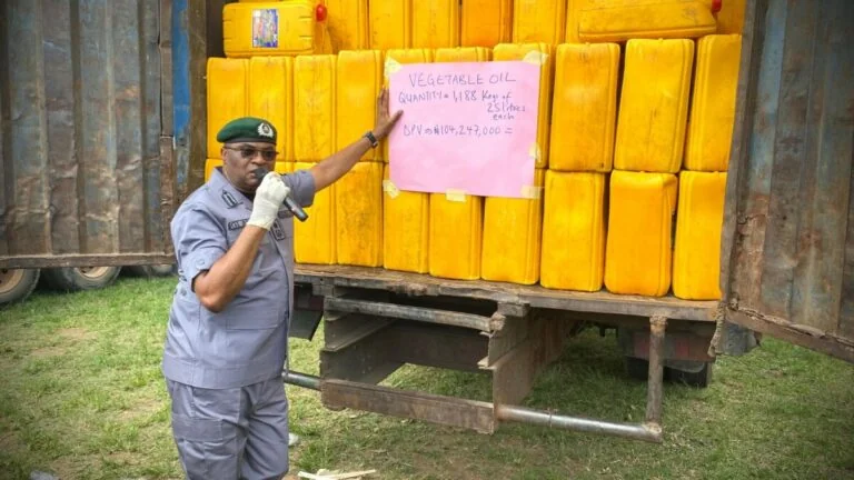 seized cartons of foreign vegetable oil - THEWILL NEWS MEDIA Uniformed man with a microphone stands beside a trailer loaded with yellow jerrycans and a pink sign about vegetable oil quantities.