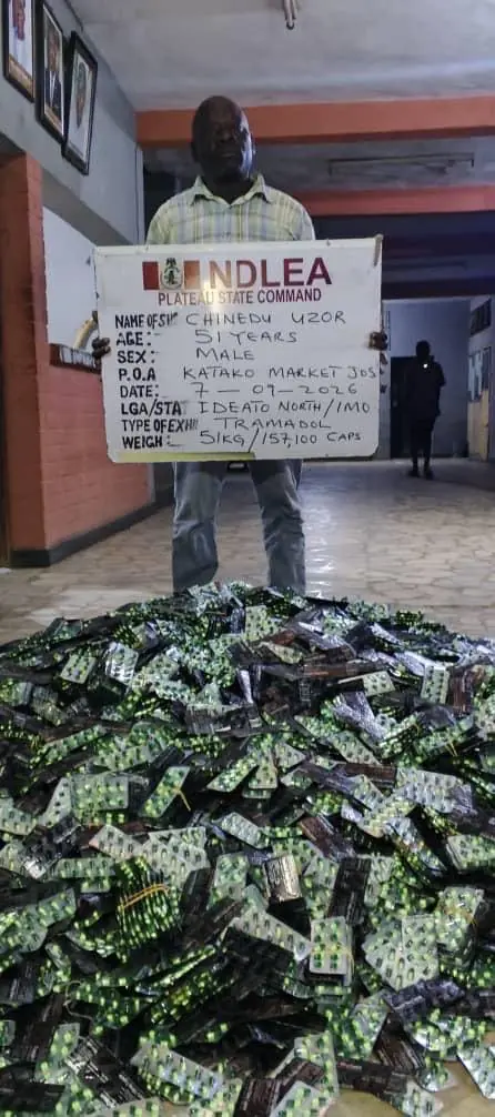 785000 Pregabalin Capsules Seized - THEWILL NEWS MEDIA Man wearing a plaid shirt stands in a hallway holding a white sign that reads NDLEA Plateau State Command, with a large pile of seized drug blister packs on the floor in front of him.