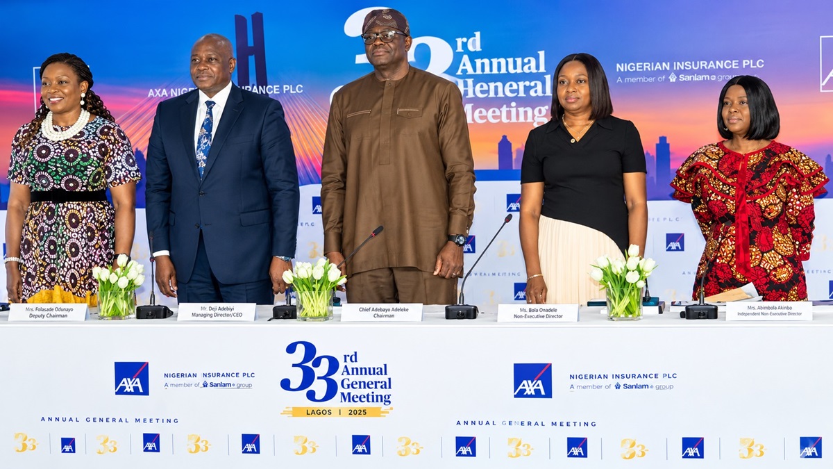 AXA Mansard AGM - THEWILL NEWS MEDIA Five professionals stand behind a long white conference table on a stage, with flower arrangements and nameplates, at the 33rd Annual General Meeting in Lagos 2025.