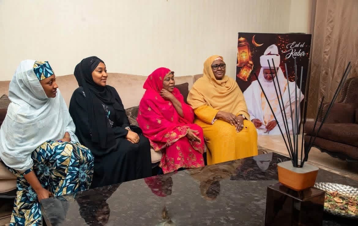 Aisha Buhari Pay Atiku visit - THEWILL NEWS MEDIA Four women wearing colorful headscarves sit on a beige sofa in a living room, chatting beside a large poster.