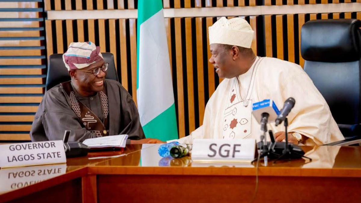 Babajide Sanwo-Olu and SGF Senator George Akume - THEWILL NEWS MEDIA Two Nigerian officials in traditional attire chat and smile at a conference table, with a Lagos State flag visible in the background and an SGF nameplate on the table nearby.