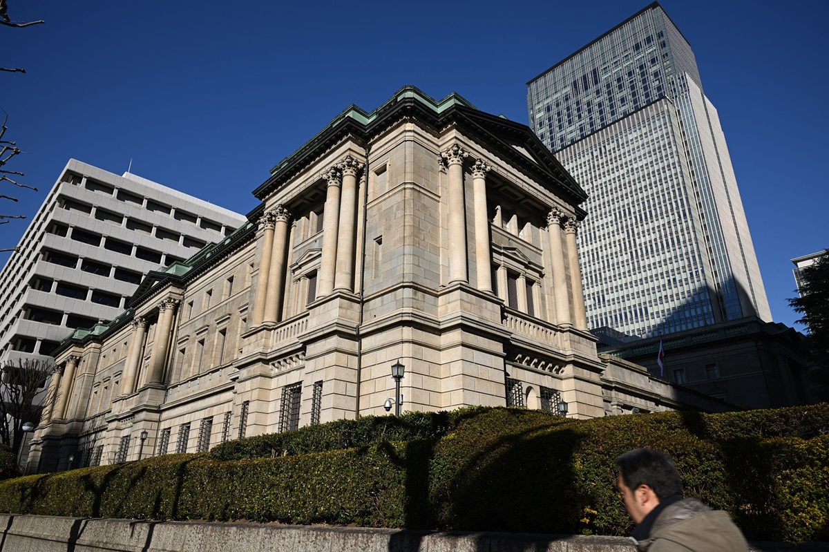 Bank of Japan headquarters building - THEWILL NEWS MEDIA Historic stone government building with tall columns beside a modern glass skyscraper under a bright blue sky; a passerby appears in the foreground.