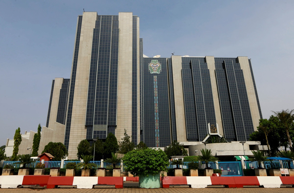 Central Bank of Nigeria CBN - THEWILL NEWS MEDIA Large modern office building with a tall glass facade, crest emblem on the center, and landscaped foreground with a red‑and‑white barrier.