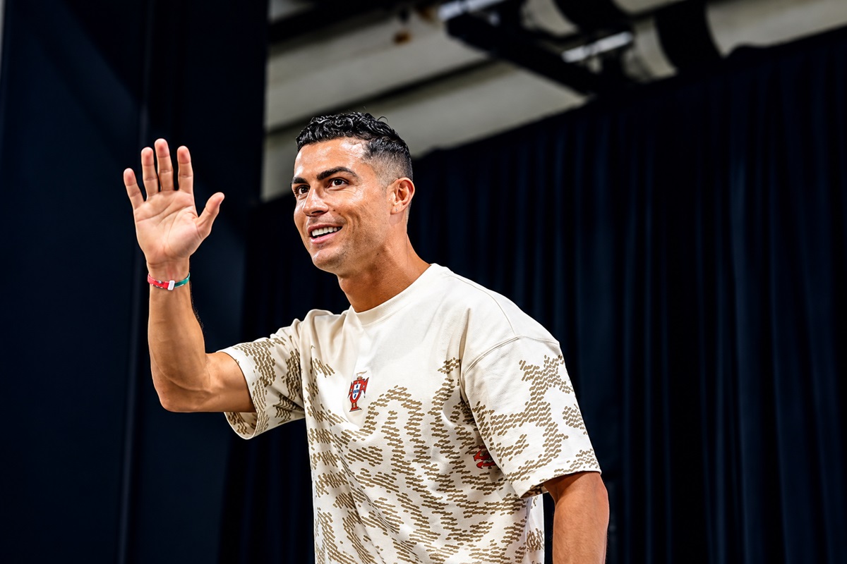Cristiano Ronaldo - THEWILL NEWS MEDIA Athlete wearing a beige patterned T-shirt waves hello from a stage with dark curtains in the background.