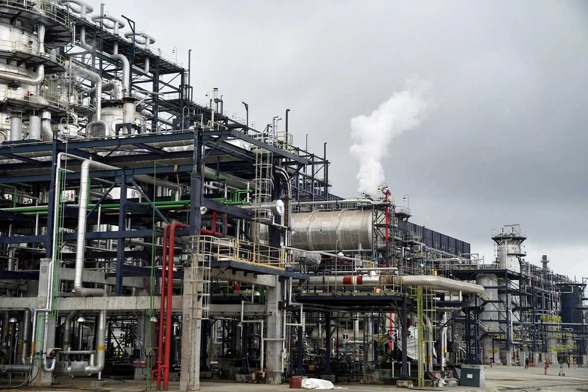 Dangote Refinery - THEWILL NEWS MEDIA Industrial refinery with dense network of metal pipes and platforms; steam plume rising from a chimney in an overcast sky.