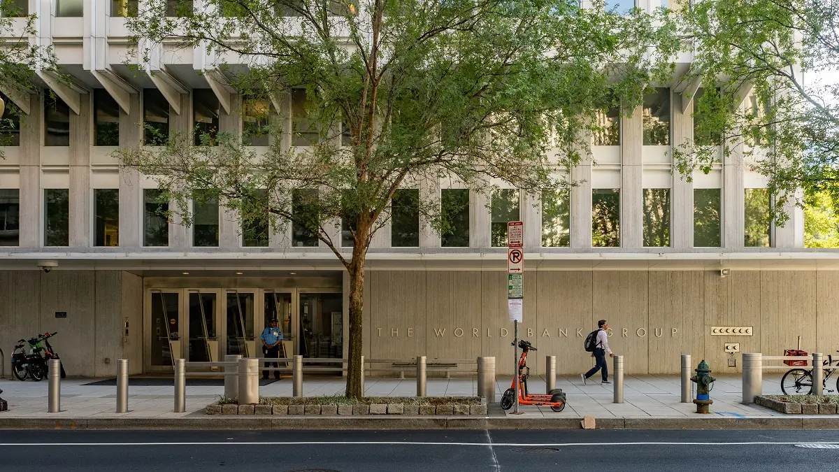 World Bank - THEWILL NEWS MEDIA Facade of The World Bank Group building with a tree in front, pedestrians nearby, and an orange scooter on the sidewalk.