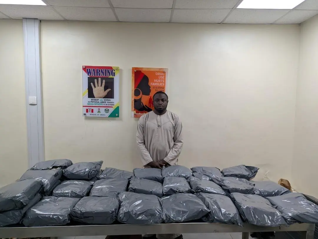 Man standing behind a stainless table covered with many gray package bundles in a room with warning posters on the wall.
