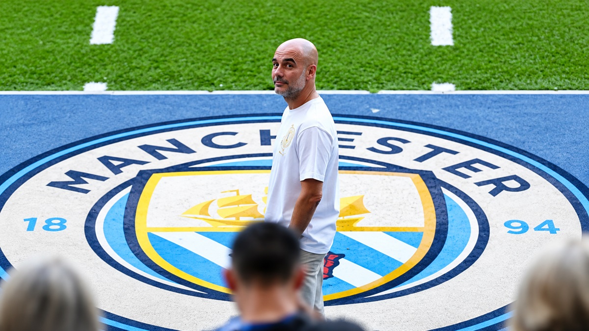 Pep Guardiola - THEWILL NEWS MEDIA Man wearing a white shirt stands on a large Manchester City emblem painted on the field with green turf behind him.