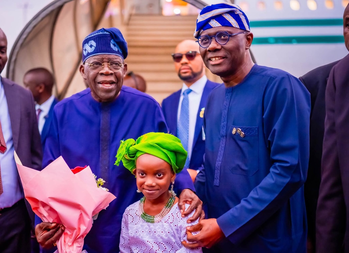 Tinubu and Sanwo Olu - THEWILL NEWS MEDIA Group of people in matching blue traditional outfits posing with a smiling young girl in a white dress and bright green headwrap holding a pink bouquet in front of an airport backdrop.