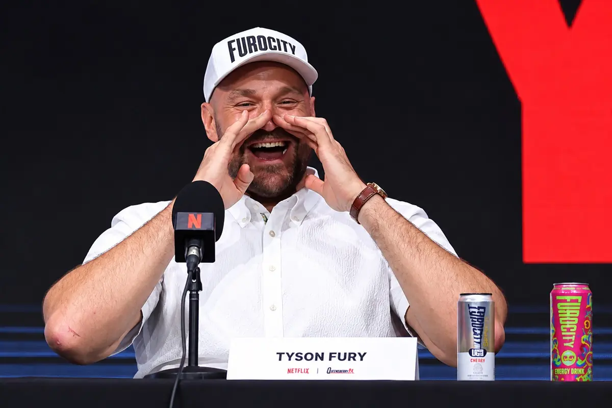 Tyson Fury - THEWILL NEWS MEDIA A male boxer at a press conference, laughing with hands cupped around his mouth; white shirt and cap with bold text, microphone in front.