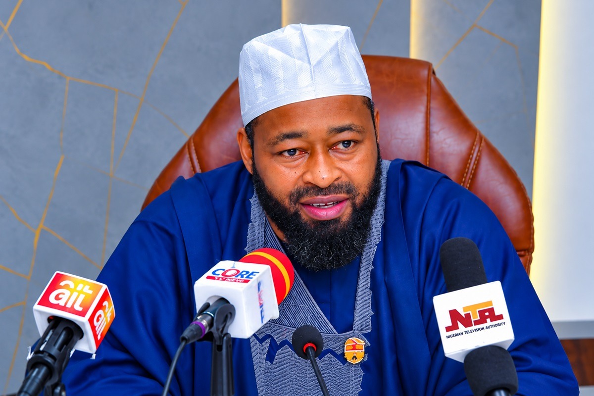 Umar Bago - THEWILL NEWS MEDIA Man in a blue traditional agbada and white cap speaks at a press conference with multiple microphones in front of him.