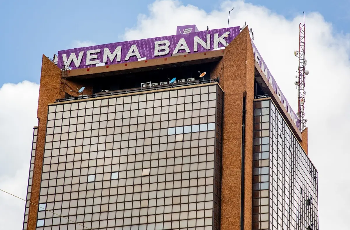 WEMA BANK - THEWILL NEWS MEDIA Tall office building with a purple WEMA BANK sign atop, against a partly cloudy sky.