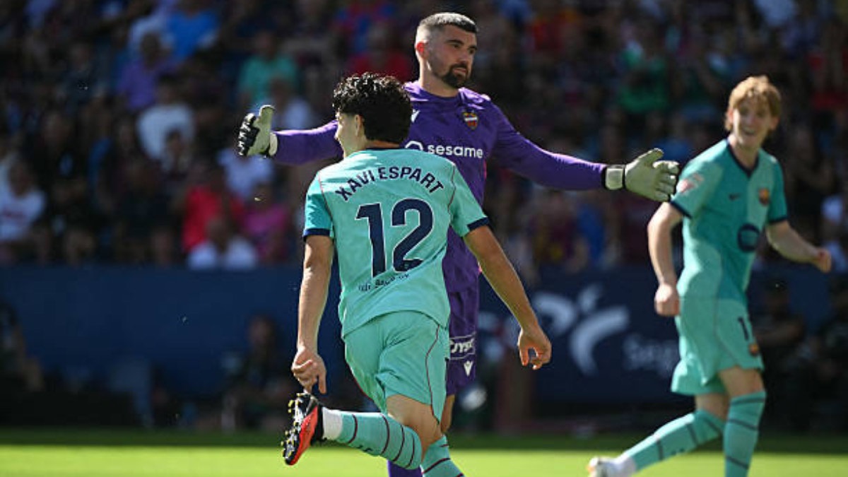 Xavi Espart celebrating his first goal for Barcelona - THEWILL NEWS MEDIA Teal-clad soccer player wearing number 12 runs toward the ball while a purple-clad goalkeeper stretches his arms in a defensive stance on a sunny field with spectators in the background.