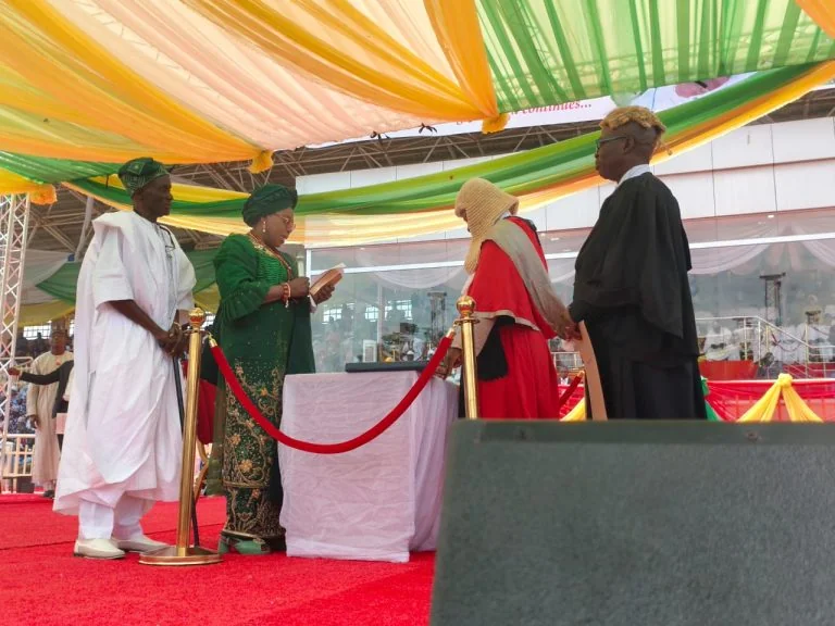 Oyebanji sworn in as Ekiti governor for second term - THEWILL NEWS MEDIA Five people on a red-carpet stage under colorful canopies, wearing traditional and academic robes, gathered around a table with a red rope barrier for a formal ceremony.