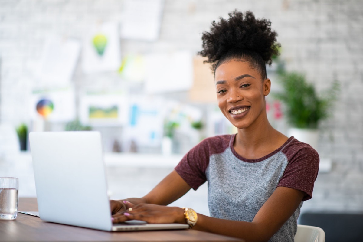 Applicants - THEWILL NEWS MEDIA Smiling woman typing on a laptop at a desk in a bright office.