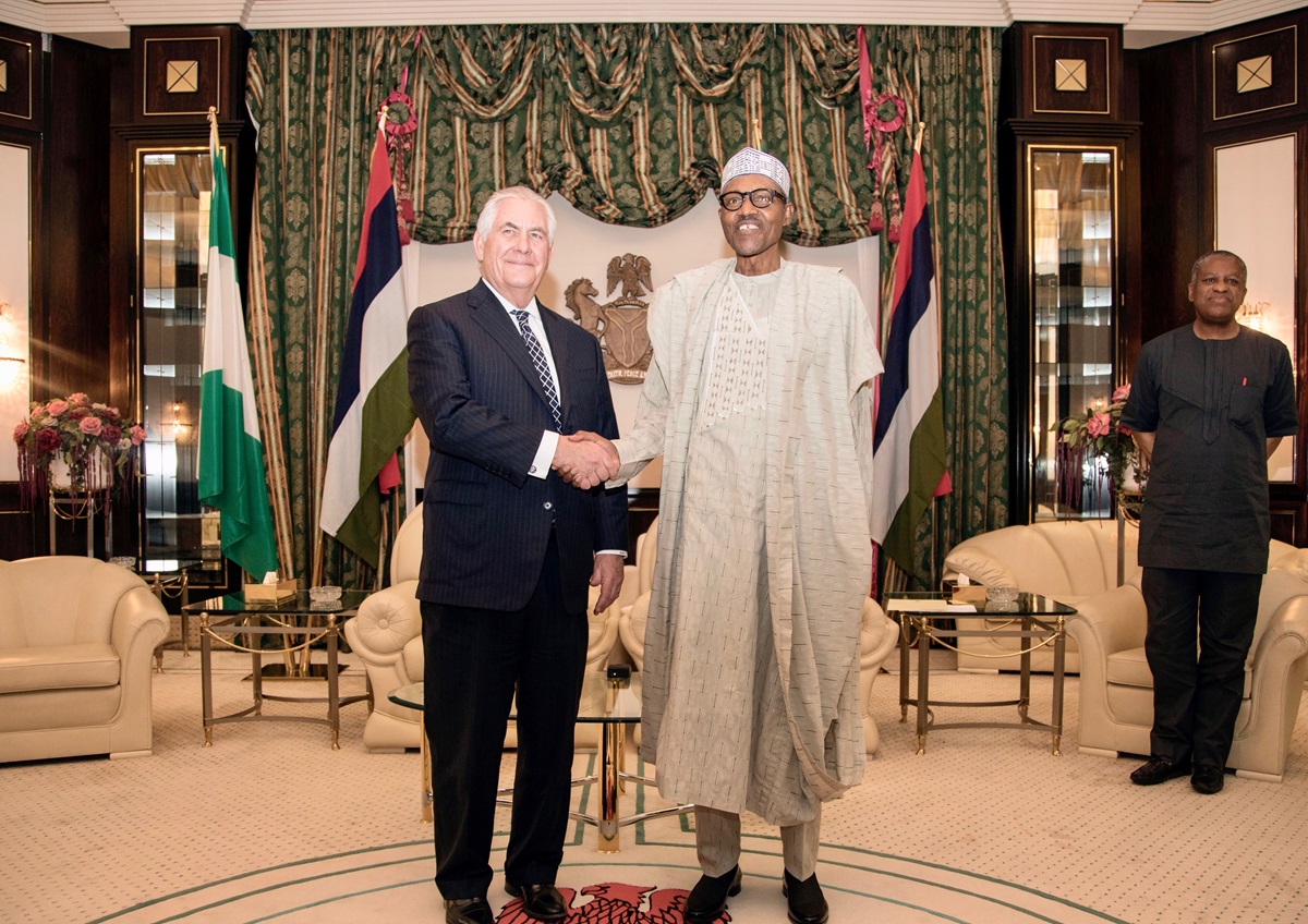 Buhari - THEWILL NEWS MEDIA Two men shake hands in a formal room with green curtains and flags behind them; one in a dark suit, the other in a light traditional Nigerian outfit. A staff member stands to the right.