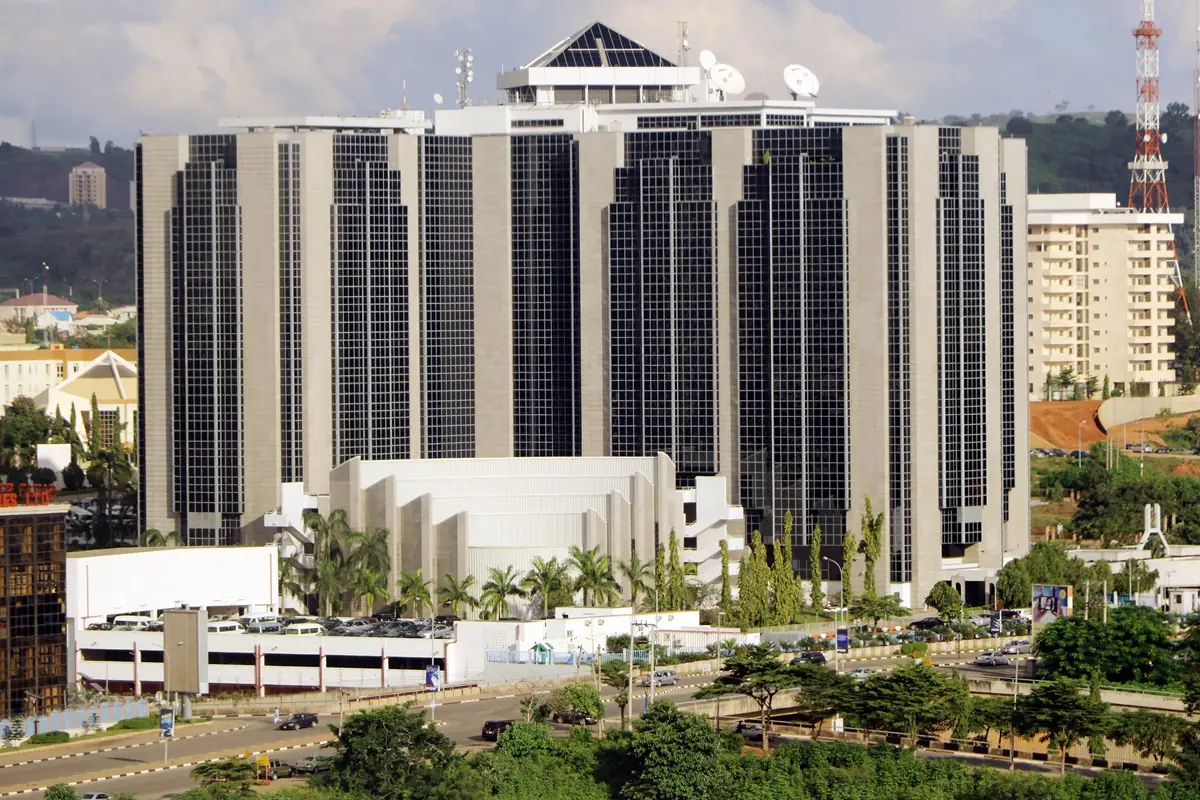 CBN - THEWILL NEWS MEDIA Large modern high-rise building with dark glass windows and a white podium, surrounded by palm trees and a busy street.