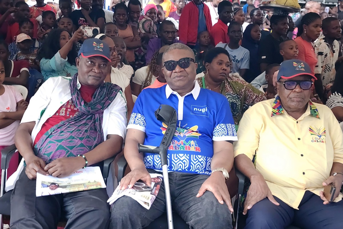 Carnival Calabar - THEWILL NEWS MEDIA Three men sit in front row at a crowded outdoor event, one wearing a blue NUGI shirt and sunglasses, flanked by two men in patterned shirts and hats.