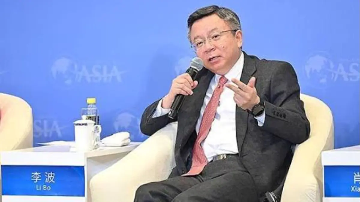 Director Bo Li - THEWILL NEWS MEDIA A man in a dark suit and pink tie sits in a cream chair, holding a microphone and gesturing during a panel discussion against a blue backdrop.
