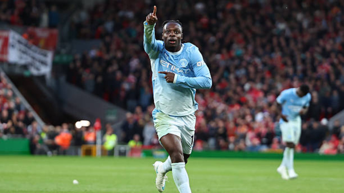 Doku - THEWILL NEWS MEDIA Soccer player in a light blue Manchester City kit points upward in celebration on the pitch with a cheering crowd behind him.