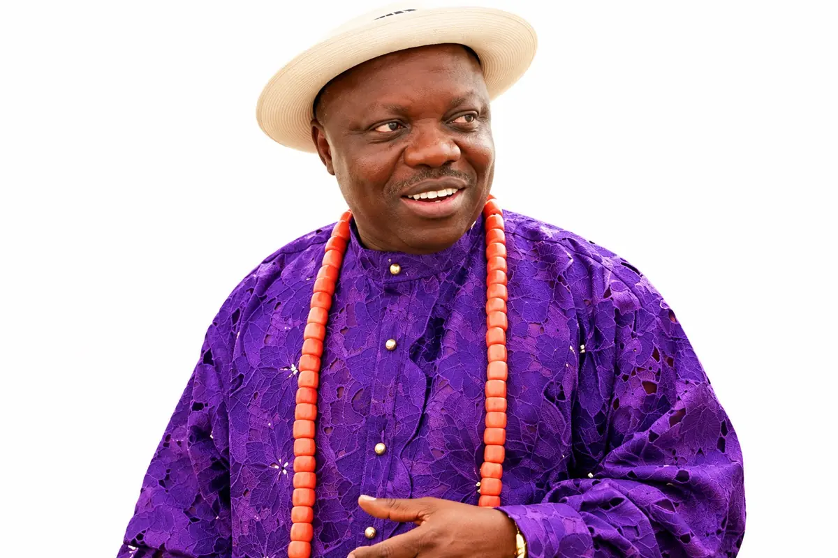 Dr Emmanuel Eweta Uduaghan - THEWILL NEWS MEDIA Man wearing a purple traditional outfit and an orange bead necklace, white hat, smiling slightly against a white background.