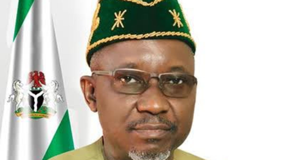 Ekpo - THEWILL NEWS MEDIA Head-and-shoulders portrait of a man wearing a green and gold traditional hat and glasses, with a Nigerian flag in the background.