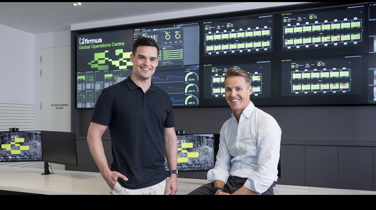 Firmus executives - THEWILL NEWS MEDIA Two men pose in a modern control room with multiple dashboard screens showing graphs and metrics in the background.