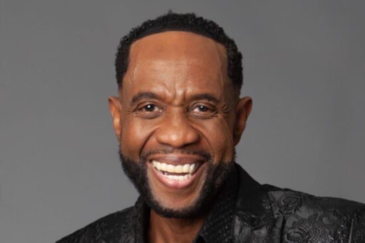 Freddie Jackson - THEWILL NEWS MEDIA Head-and-shoulders portrait of a smiling Black man with a short beard and curly hair, wearing a black patterned blazer against a gray background.