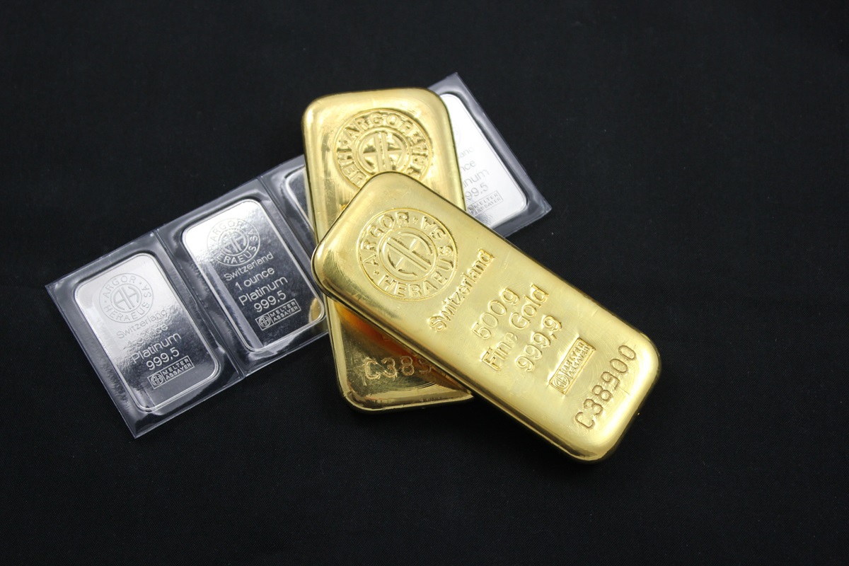 Gold and platinum bullion bars - THEWILL NEWS MEDIA A small pile of gold bars with one large gold ingot on top, inscribed with 500 g fine gold 999.9 and assay marks, resting on a dark surface with silver bars nearby.