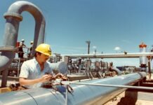 Worker in a yellow hard hat repairing electrical wiring on large stainless steel pipelines at an industrial refinery site.