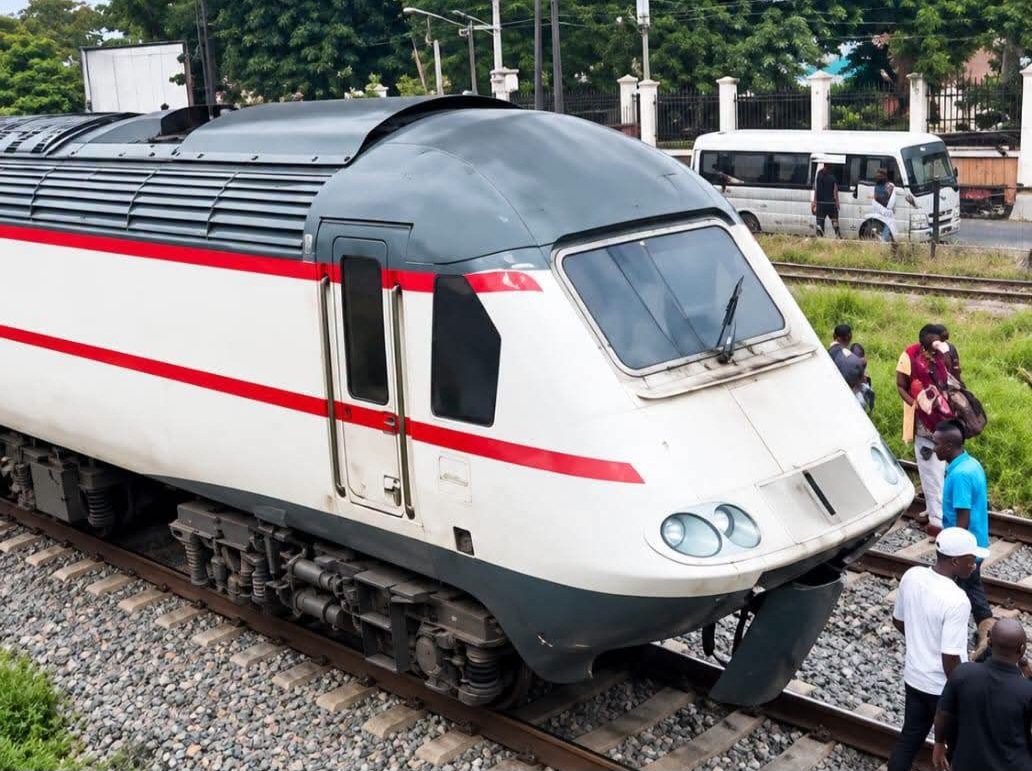 Lagos Train - THEWILL NEWS MEDIA Front of a white passenger train with a red stripe on the nose, parked on tracks while several people stand nearby on the platform side.