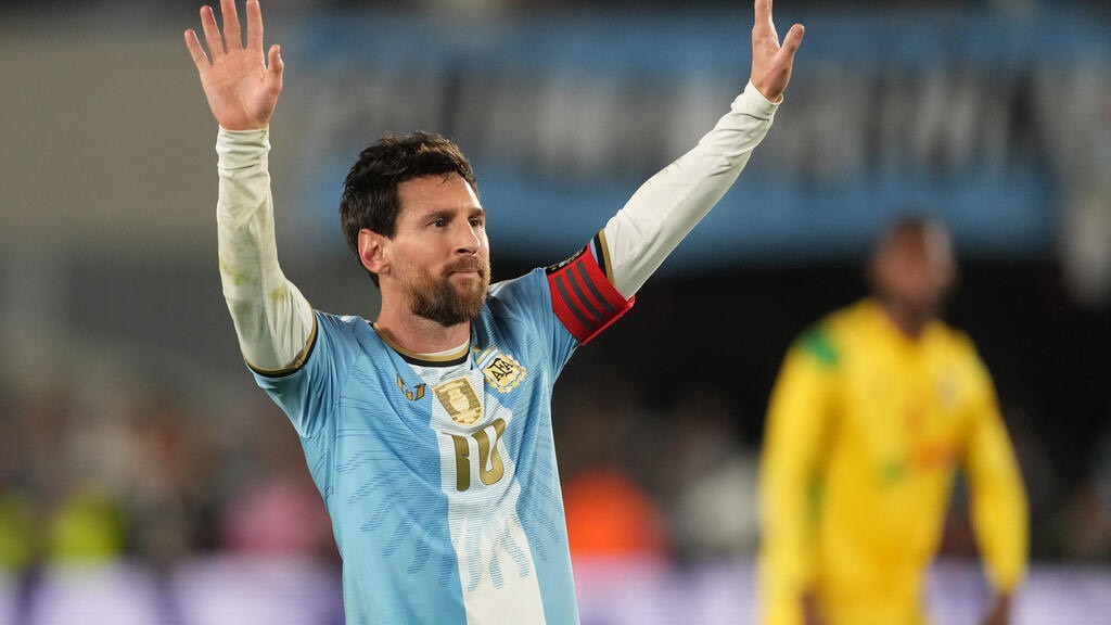 Lionel Messi - THEWILL NEWS MEDIA Argentina national team captain raises both arms in celebration on the pitch, wearing a light blue jersey with the number 10 and a red armband.
