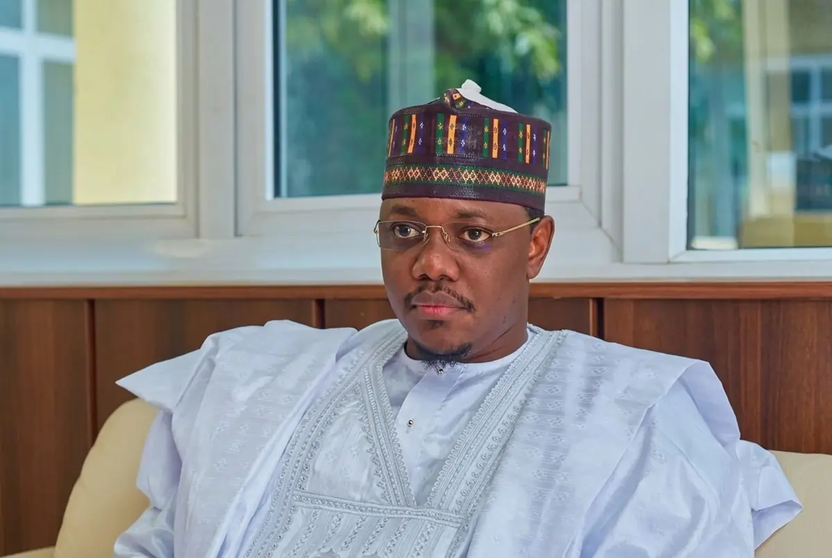 Mustapha Sule Lamido - THEWILL NEWS MEDIA Man in a white traditional outfit with a colorful patterned cap seated on a sofa by a window, looking slightly to the left.